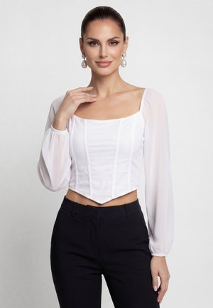 Žena s tmavými vlasy v bílé blůze s čtvercovým výstřihem a průsvitnými dlouhými rukávy a černými kalhotami, nosící velké perlové náušnice s visacím designem.