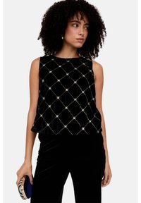 Schwarzes ärmelloses Samttop mit einem Diamantmuster und dekorativen Akzenten. Kombiniert mit einer schwarzen Hose und einer Clutch in der Hand.