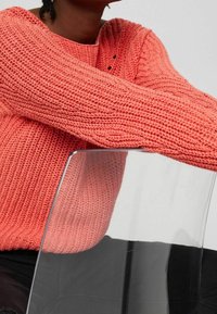 Korale sweter dziewiarski z fakturowanym wzorem i okrągłym dekoltem, w zestawieniu z przezroczystym akrylowym krzesłem. Zbliżenie na detale materiału.