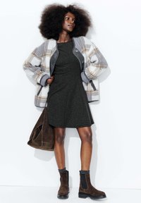 Veste en peluche à carreaux gris et crème, robe noire texturée, bottes à cheville en daim marron et sac à épaule marron, sur fond blanc.