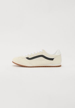 Baskets beige avec des lacets blancs, une bande noire sur le côté, un détail blanc au talon et une semelle en caoutchouc marron, présentées sur un fond blanc uni.