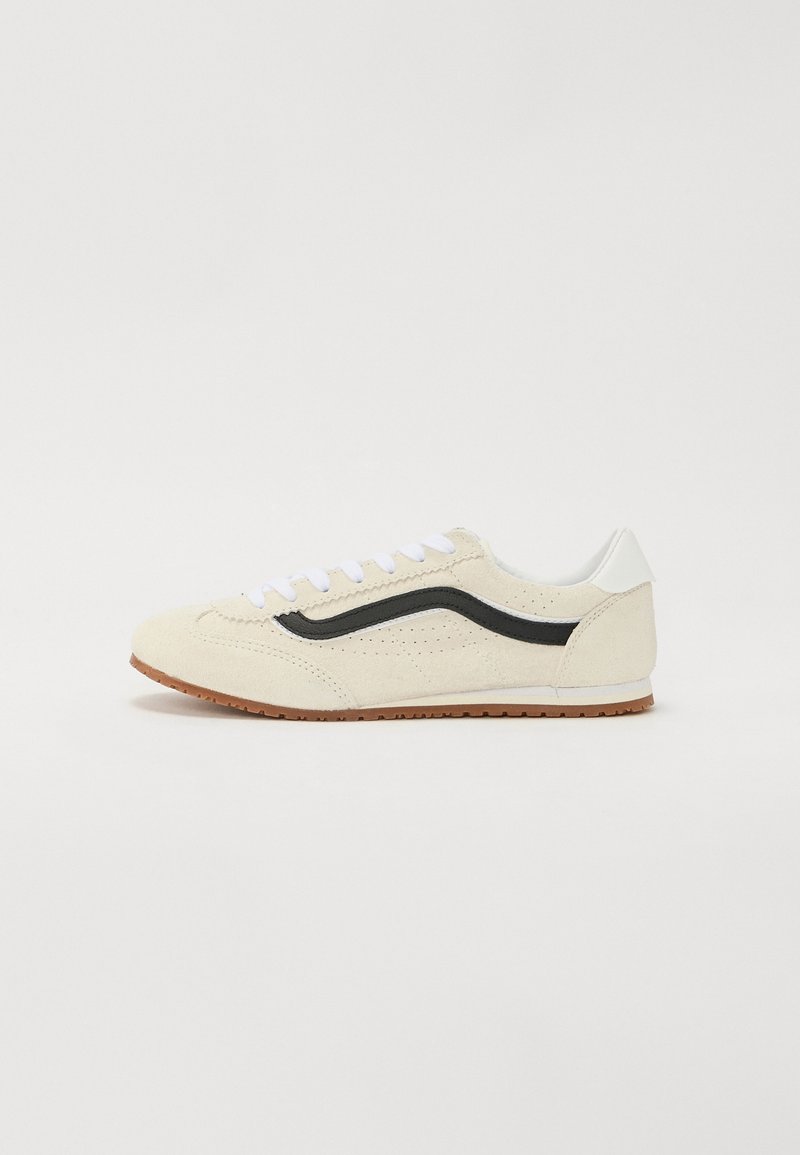 Baskets beige avec des lacets blancs, une bande noire sur le côté, un détail blanc au talon et une semelle en caoutchouc marron, présentées sur un fond blanc uni.