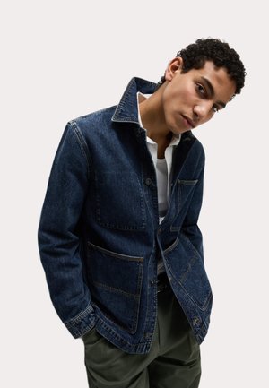 Chaqueta de mezclilla azul oscura con cuello, cuatro bolsillos frontales y detalles de costura visibles. Lleva puesta una camisa blanca.