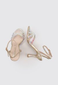 Sandalias de tacón alto con tiras, con un acabado metálico dorado, adornadas con coloridos strass en la tira del dedo, suela plana y correa ajustable en el tobillo.