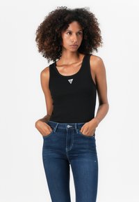 Débardeur noir avec un col arrondi, en coton, arborant un petit logo triangle argenté sur la poitrine. Associé à un jean bleu taille haute.