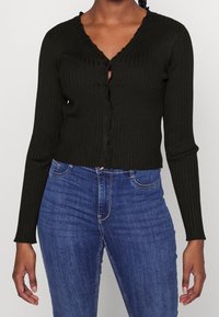 Cardigan nero a costine con scollo a V, maniche lunghe e bordi scallop; presenta chiusura con bottoni e design corto. Abbinato a jeans blu.