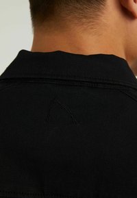 Gros plan sur le cou et le haut du dos d'une personne portant une chemise noire texturée avec un logo brodé en relief près du col.