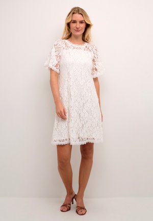 Weißes Spitzenkleid mit kurzen, schwingenden Ärmeln und einem gewellten Saum. Der Stoff zeigt ein filigranes Blumenmuster. Getragen mit braunen Sandalen mit Absatz.