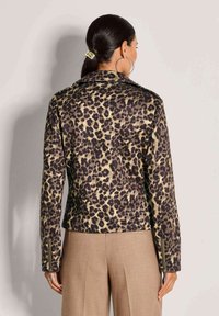 Vrouw met donker haar in een paardenstaart, die een jas met luipaardprint en beige broek draagt, gezien vanaf de achterkant tegen een effen lichte achtergrond.