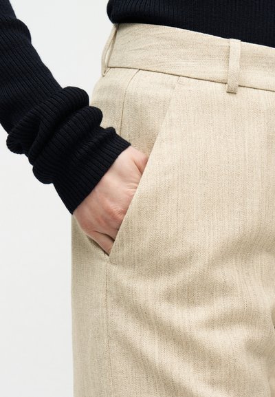 Hånd plassert i frontlomme på beige teksturerte bukser, brukt med svart ribbet langermet topp.