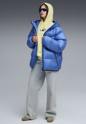 Giacca puffa blu con cappuccio e bordi neri, indossata sopra un felpa gialla. Abbinata a jeans a gamba larga chiari e scarpe grigie.