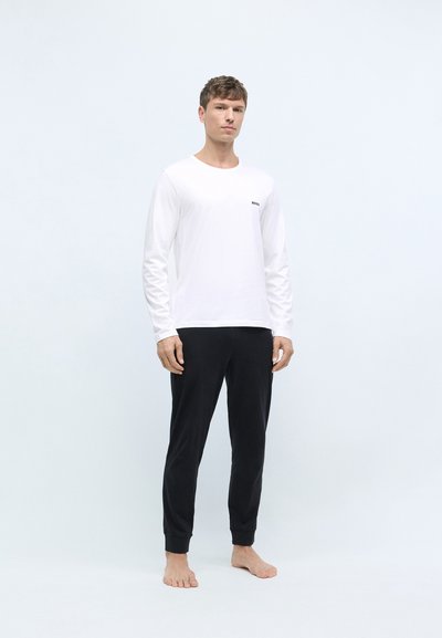 Chemise blanche à manches longues avec un petit logo noir, associée à un pantalon de détente noir et ajusté en tissu doux. Modèle se tenant pieds nus.