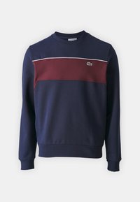 Felpa navy con pannello frontale bordeaux, striscia bianca a contrasto e logo Lacoste. Presenta un collo rotondo e polsini e orlo a costine.
