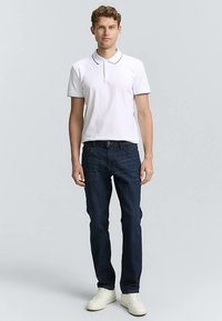 Weißes Poloshirt mit Kragen, kombiniert mit dunkelblauen Jeans. Das Outfit wird durch weiße Sneakers ergänzt. Einfaches, klassisches Design mit tailliertem Schnitt.