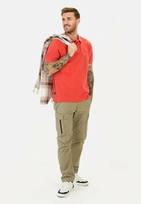 Rød polo skjorte med korte ærmer og et lille logo, parret med beige cargo bukser med lommer og sorte lynlåse. Hvide sneakers.