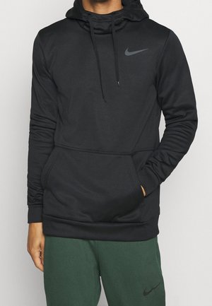 Mężczyzna ubrany w czarną bluzę Nike z kapturem i przednią kieszenią oraz zielone spodnie dresowe Nike, stojący na jasnym, jednolitym tle.