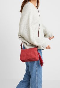 Femme portant un pull gris clair et un jean bleu, portant un sac bandoulière rouge texturé avec un détail à frange.