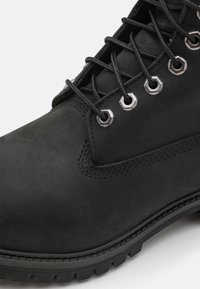 Botas de cuero negro con superficie texturizada, ojales metálicos hexagonales y cordones negros. La suela de goma cuenta con relieves profundos para mayor tracción.