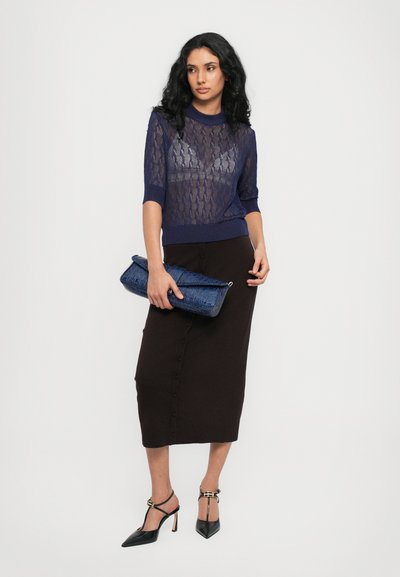Suéter de punto azul marino de manga corta, superpuesto a una falda midi marrón con botones frontales; sosteniendo un bolso texturizado azul; zapatos de tacón negros.