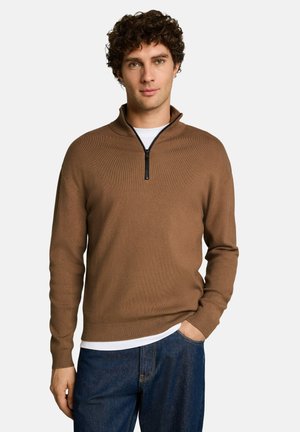 Uomo con capelli ricci che indossa un maglione marrone con mezza cerniera sopra una camicia bianca e jeans blu, in piedi con una mano in tasca.