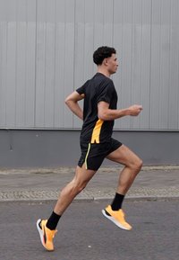 Maglietta sportiva nera con accenti arancioni, pantaloni corti neri e scarpe da corsa arancioni. Il soggetto è in movimento su una superficie asfaltata.