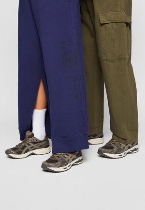 Long navy blue pants with a side slit over an olive green pair, featuring pockets and textured sneakers in shades of brown and gray.
