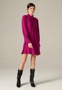 Abito magenta a maniche lunghe con colletto alto, parte anteriore plissettata, maniche svasate e orlo sopra il ginocchio, abbinato a stivali neri fino al ginocchio.