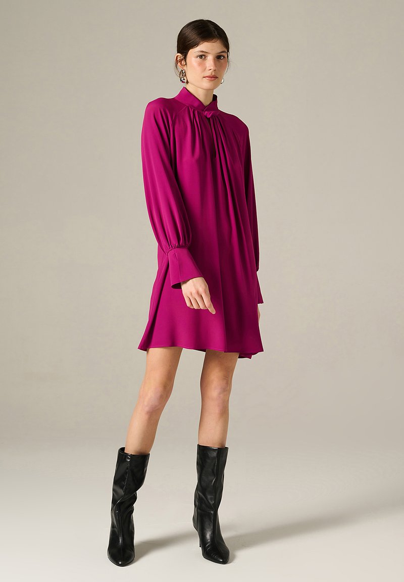 Abito magenta a maniche lunghe con colletto alto, parte anteriore plissettata, maniche svasate e orlo sopra il ginocchio, abbinato a stivali neri fino al ginocchio.