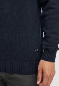Navy knitted sweater with ribbed cuffs and hem, featuring a small fabric label. The texture is smooth and stretchy, providing a comfortable fit.