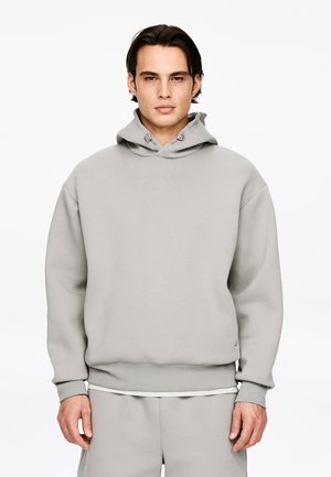 Młody mężczyzna ubrany w jasnoszarą bluzę z kapturem i pasujące spodnie, stojący na tle jednolitego białego tła.