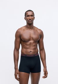 Muscular man standing against plain background wearing black Björn Borg boxer briefs, looking forward with a neutral expression.