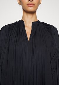Černá plisovaná blůza s výstřihem do V. Tkanina má hladkou strukturu s vertikálními záhyby, což dodává strukturovaný vzhled.