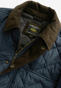 Steppjacke mit einem navyblauen Äußeren, braunem Cordkragen, metallischem Reißverschluss und einem Etikett mit der Aufschrift "Premium-Qualität, langlebige Waren."