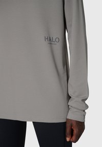 Långärmad, ljusgrå träningsskjorta gjord av texturerat, fuktavvisande tyg. Har "HALO"-logotyp i mörka bokstäver nära fållen.