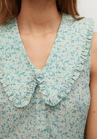 Kukallinen napitettava paita, jossa on röyhelökaulus; pehmeää kangasta vaaleansinisenä, valkoisilla ja tummansinisillä kuvioilla; V-pääkkausmuoto.