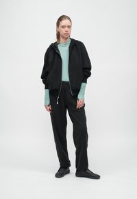 Μαύρο φούτερ με φερμουάρ και χαλαρή εφαρμογή, φορεμένο πάνω από ένα πετρόλ μακρυμάνικο μπλουζάκι και συνδυασμένο με μαύρο παντελόνι και αθλητικά παπούτσια. Μινιμαλιστικός σχεδιασμός.