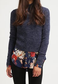 Pull en maille côtelée marine avec un col haut, superposé sur une chemise à imprimé floral avec des fleurs rouges et crème sur un fond sombre.
