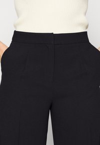 Zbliżenie na czarne, dopasowane spodnie z bocznymi kieszeniami, noszone z wpuszczonym w spodnie sweterkiem w kolorze kremowym o prążkowanej fakturze.