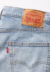 Lichtblauwe denim jeans met een leren label met rode en zwarte graphics, vijf zakken ontwerp en kenmerkende gele stiksels.