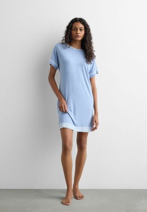 Jeune femme aux cheveux bouclés, debout pieds nus, portant une robe bleu clair à manches courtes avec un galon en dentelle blanche aux manches et à l'ourlet.