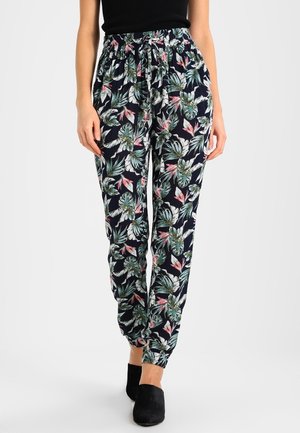 Femme portant un pantalon noir fluide à imprimé floral, avec taille et poignets élastiques, associé à des chaussures slip-on noires et un haut noir.
