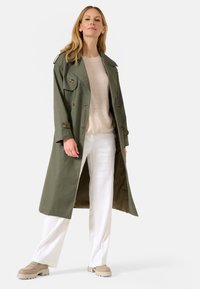 Manteau trench vert olive avec des boutons double boutonnage et des épaulettes, porté sur un haut beige et un pantalon blanc. Présente des manches longues et un design fluide.