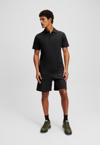 Cămașă polo neagră cu mâneci scurte și guler, asortată cu pantaloni scurți negri și adidași verzi. Textură netedă, branding minim.