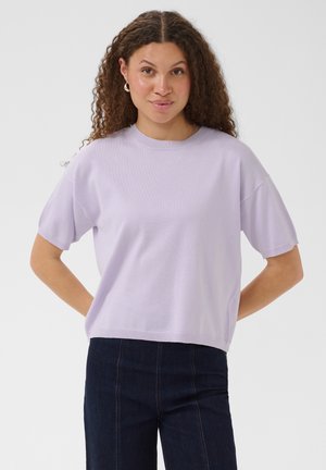 Femme aux cheveux bouclés portant un pull à manches courtes violet clair et un pantalon taille haute bleu foncé sur un fond uni.