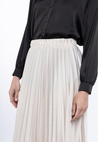 Cămașă neagră din satin, cu nasturi, purtată cu o fustă plisată crem, dotată cu un brâu elastic, evidențiind textură netedă și material fluid.
