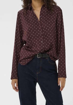 Femme portant une chemise à manches longues marron foncé avec des pois rose clair, rentrée dans un jean bleu foncé avec une ceinture noire.