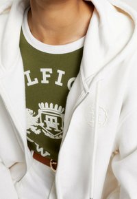 Sudadera blanca con cremallera hecha de una tela suave y con un logo bordado. Lleva encima una camiseta verde oliva que presenta texto blanco y un diseño gráfico.
