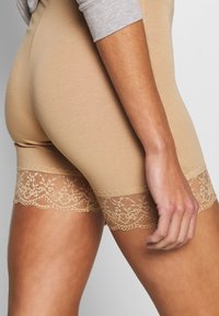 Kraťasy v tělové barvě z měkké tkaniny, s krajkovou ozdobou na lemu zdobenou květinovými vzory, která vytváří texturovaný kontrast.