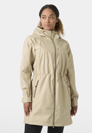 Mujer con un impermeable beige con capucha, cremallera frontal y bolsillos con cremallera, de pie contra un fondo claro y sencillo.
