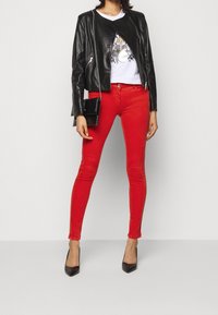 Chaqueta de cuero negra con diseño asimétrico, camiseta blanca gráfica, jeans skinny rojos y tacones altos negros. Pequeño bolso negro con tachuelas.